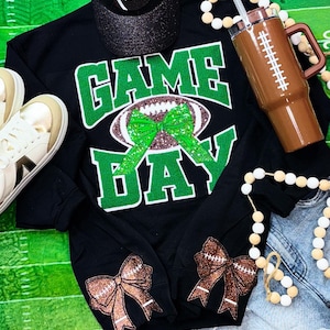 May include: Black sweatshirt with green glitter text that says "GAME DAY" with a football and a green bow. The sleeves have brown glitter bows with football stitching.