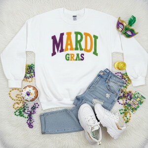 May include: White sweatshirt with the text "MARDI GRAS" in purple, orange, and green. The image includes denim jeans, white trainers, a decorative mask, and Mardi Gras beads. The sweatshirt is a festive apparel item.