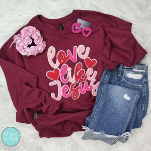 Peut inclure: Sweat-shirt bordeaux avec l'inscription "Love Like Jesus" en lettres roses et rouges. L'image comprend un short en jean bleu, un chouchou rose et des boucles d'oreilles en forme de cœur.