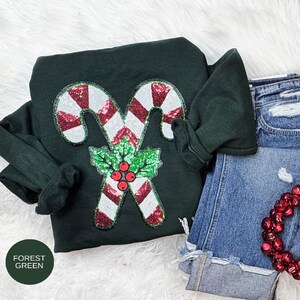 May include: Forest green sweatshirt with a sequined candy cane design. The candy canes are red and white, with a green holly and red berry accent. The sweatshirt is paired with denim shorts and a red beaded garland.