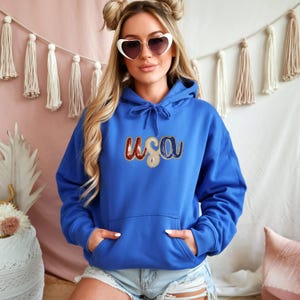 May include: A royal blue hoodie with the word "USA" embroidered in red, white, and blue. The hoodie has a drawstring hood and a front pocket. The image is of a person wearing the hoodie.