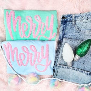 May include: Two pastel-colored sweatshirts, one mint green and one light blue, each embroidered with the word "Merry" in pink. A pair of denim shorts with rhinestone embellishments and two decorative light bulbs are also present. A garland with colorful pom-poms is at the bottom.
