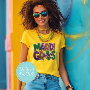 May include: A yellow t-shirt with the words "Mardi Gras" in green, purple, and gold glitter. The shirt is paired with blue jeans, sunglasses, and colourful jewellery. The Sweet Tee Y'all logo is in the bottom left corner.