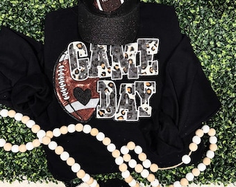 Può includere: Felpa nera con grafica di football americano e la scritta "GAME DAY" in stampa leopardata. Un berretto da baseball nero con un disegno di football americano è sopra. Una collana di perle bianche e beige è in primo piano.