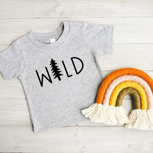 Może przedstawiać: Szary T-shirt z nadrukiem słowa "WILD" w kolorze czarnym, ze stylizowanym drzewem sosnowym zastępującym literę "I". Koszulka leży obok tęczowej makramowej wiszącej ozdoby ściennej.