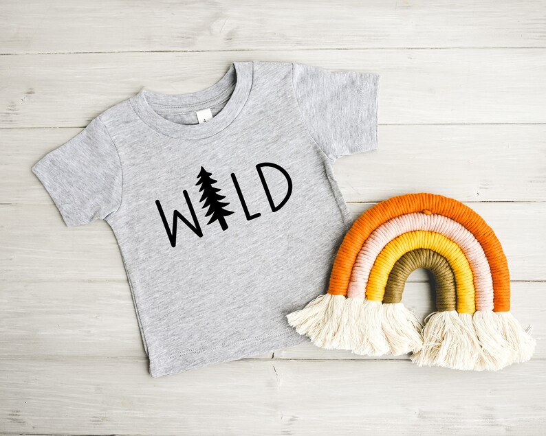 Może przedstawiać: Szary T-shirt z nadrukiem słowa "WILD" w kolorze czarnym, ze stylizowanym drzewem sosnowym zastępującym literę "I". Koszulka leży obok tęczowej makramowej wiszącej ozdoby ściennej.