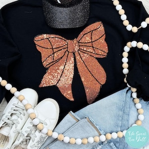 May include: Black sweatshirt with a large, sparkly, copper-colored bow design. The bow is made of sequins and has a shiny, metallic finish.