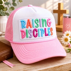 Può includere: Cappellino da camionista rosa e bianco con la scritta "RAISING DISCIPLES" ricamata a lettere colorate. Il cappellino ha una visiera rosa, un retro in rete e un pannello frontale bianco.
