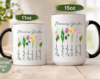 May include: Two white ceramic mugs with black handles and rims. The mugs feature the text Nannies Garden and a floral illustration with names printed below each flower. One mug is labeled 11oz and the other is labeled 15oz in green text.