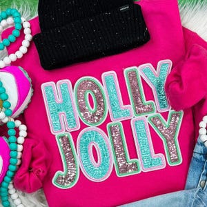 May include: A bright pink sweatshirt with the words "HOLLY JOLLY" in sequined letters. The letters are outlined in light green and filled with teal and pink sequins. A black beanie sits on top of the sweatshirt.