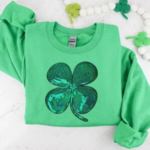 Puede incluir: Una sudadera verde con un gran diseño de trébol de cuatro hojas con lentejuelas. La camisa de manga larga es de color verde liso, con cuello redondo y puños enrollados. Prenda con temática del Día de San Patricio.