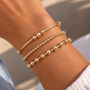 Puede incluir: Un conjunto de tres pulseras doradas en la muñeca. La pulsera superior presenta una barra lisa con dos pequeñas esferas, mientras que las pulseras del medio y la inferior consisten en pequeñas cuentas doradas uniformes unidas en un bucle.