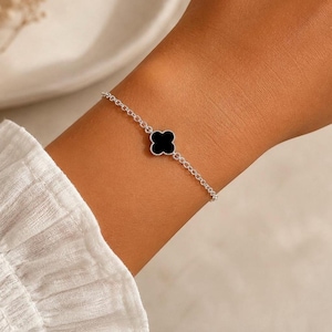 Puede incluir: Una delicada pulsera de cadena de plata que presenta un único dije de trébol de cuatro hojas centrado con una incrustación negra sólida y lisa. La pulsera está construida con eslabones de plata pulida que conectan con el motivo central.