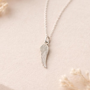 May include: A silver-colored necklace featuring a pendant shaped like a wing. The wing pendant has detailed feather etching and hangs from a delicate chain. The necklace is displayed on a light-colored surface.