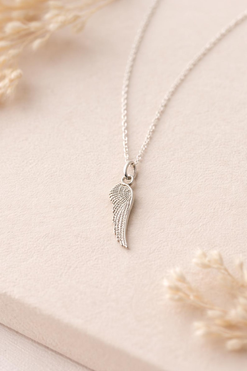 May include: A silver-colored necklace featuring a pendant shaped like a wing. The wing pendant has detailed feather etching and hangs from a delicate chain. The necklace is displayed on a light-colored surface.