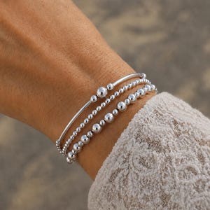 May include: A set of three silver-toned beaded bracelets worn together. The bracelets feature varying sizes of polished spherical beads and smooth cylindrical bars, creating a layered look with a reflective metallic finish on the wrist.