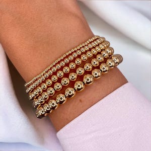 May include: Several gold beaded bracelets of different sizes are stacked on a wrist. The bracelets feature small to large spherical beads, all in a consistent gold tone. The arrangement creates a layered effect, showcasing the varying bead sizes and the overall shine.