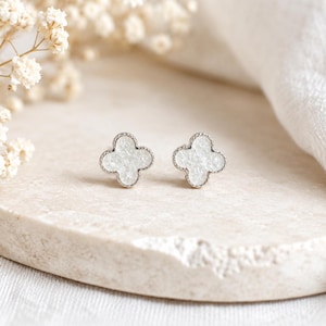 May include: Two clover-shaped stud earrings with a white, textured centre and a silver-coloured border. The earrings are presented on a light-coloured, textured surface. Soft, white flowers are visible in the background.