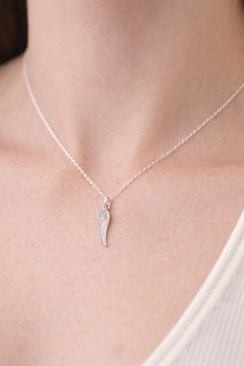 May include: A delicate silver necklace featuring a small pendant shaped like a wing. The wing pendant is detailed with fine lines, and the necklace chain is a simple, thin design. The necklace is worn against a light background.