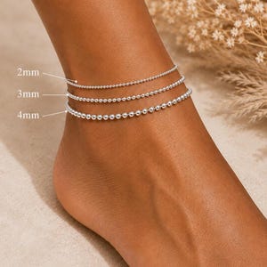 May include: Three silver-toned beaded anklets worn together, featuring spherical beads in three distinct sizes. The anklets are labelled with text indicating bead diameters of 2mm, 3mm, and 4mm, showing the variation in the size of the metallic beads.