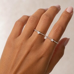 May include: Two silver beaded rings worn on adjacent fingers. One ring features a small, smooth heart-shaped charm, while the other ring features a small, smooth star-shaped charm. Both rings are constructed from a series of tiny, uniform silver spheres.