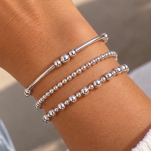 May include: A set of three silver-toned bracelets. The top bracelet features a smooth bar with three central spheres. The middle bracelet consists of small, uniform silver beads. The bottom bracelet features a mix of small and large silver beads.