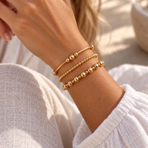 May include: A set of three gold-toned bracelets worn on a wrist. The top bracelet is a smooth bar with three small spheres, the middle is a strand of tiny uniform beads, and the bottom is a strand of larger, varying spherical gold-toned beads.