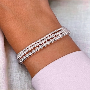 May include: Three silver beaded bracelets of different sizes are stacked on a wrist. The bracelets are made of small, round, shiny beads. The bracelets are a simple and elegant accessory.