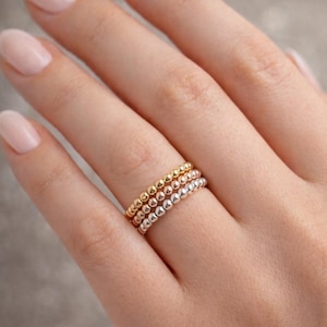 Puede incluir: Tres anillos de cuentas en oro, oro rosa y plata están apilados en un dedo. Los anillos están hechos de pequeñas cuentas redondas, creando una apariencia texturizada. El fondo es gris neutro.