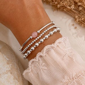 Puede incluir: Un conjunto de tres pulseras de plata. Una pulsera presenta una única gema rosa, mientras que las otras dos están adornadas con pequeñas cuentas redondas de plata. Las pulseras se apilan en una muñeca, con una manga rosa claro en el fondo.
