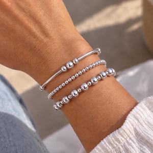 May include: A set of three silver-toned beaded bracelets worn together. The top bracelet features a smooth bar with three central beads. The middle bracelet consists of small uniform beads. The bottom bracelet features larger, varied-sized beads.