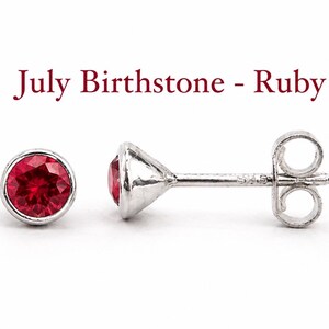 May include: A pair of silver stud earrings featuring round, faceted red ruby stones in a bezel setting. The text "July Birthstone - Ruby" is displayed above the earrings. The posts have butterfly backs and are marked with the number 925.