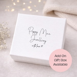 May include: A white textured gift box with the text "Poppy Mae Jewellery with love" in black script. A pink circle with the text "Add On Gift Box Available" is in the lower right corner. The background is a soft, blurred setting.
