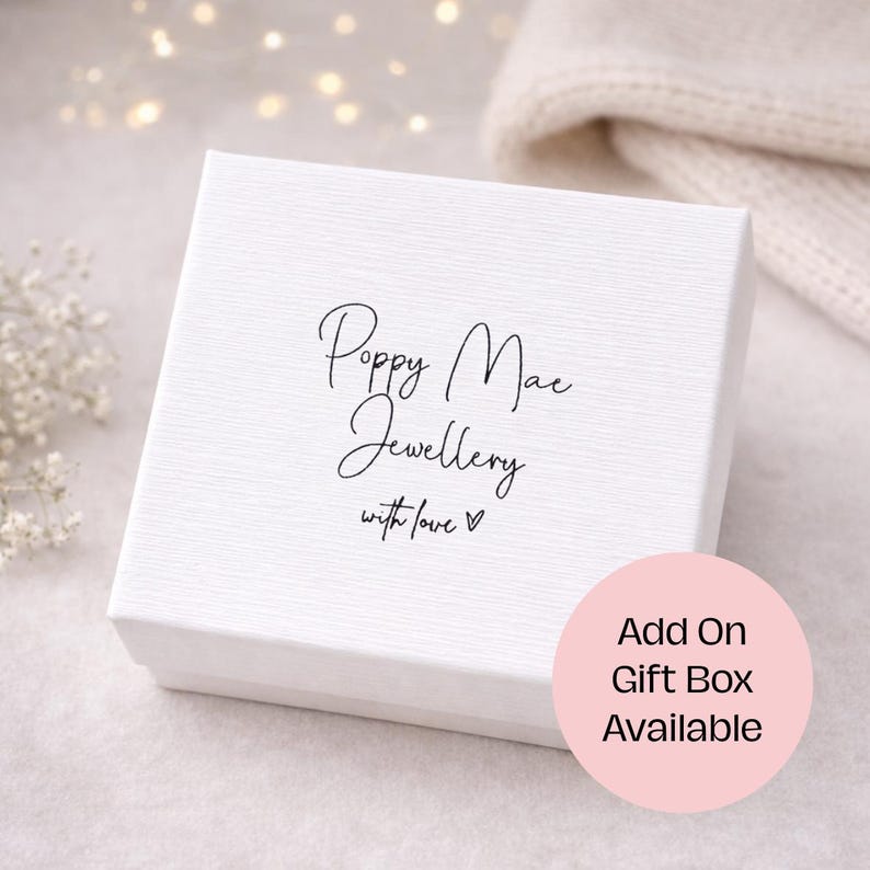 May include: A white textured gift box with the text "Poppy Mae Jewellery with love" in black script. A pink circle with the text "Add On Gift Box Available" is in the lower right corner. The background is a soft, blurred setting.