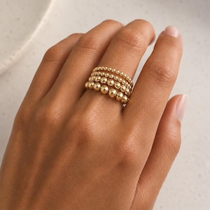 May include: A stack of four gold-toned rings, each featuring a continuous band of polished spherical beads. The rings are arranged in order of bead size, with the smallest beads at the top and the largest beads at the base of the finger stack.