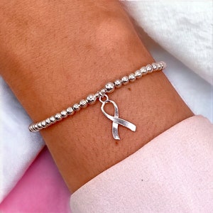 Peut inclure: Bracelet en argent avec un charm en forme de ruban. Le bracelet est composé de petites perles rondes en argent. Une perle plus grosse se trouve près du charm en forme de ruban, symbole de sensibilisation.