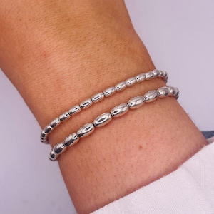 May include: Two silver beaded bracelets stacked on a person's wrist. The bracelets are made of oval-shaped beads.