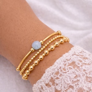 May include: Three gold bracelets are stacked on a wrist. The top bracelet has a light blue stone. The other two bracelets are made of small gold beads. The wrist is against a white background.