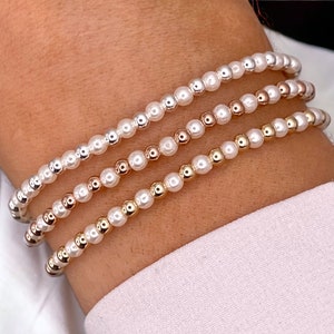 May include: Three bracelets with white pearl-like beads and gold and rose gold accents. The bracelets are stacked on a person's wrist.