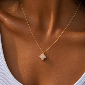 May include: A gold chain necklace featuring a four-leaf clover pendant with a textured, light-colored stone center. The pendant is framed in a thin gold border and hangs from a delicate, fine-link gold chain that rests against the collarbone.