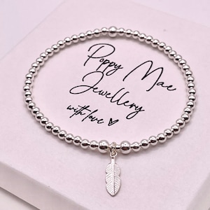 May include: A silver beaded bracelet with a silver feather charm. The bracelet is in a white box with the text "Poppy Mae Jewellery with love" written on it.