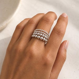 May include: A stack of four silver rings worn on a finger. Each ring features a continuous band of polished spherical beads. The beads on each ring vary in size, with the top ring having the smallest beads and the bottom ring having the largest.