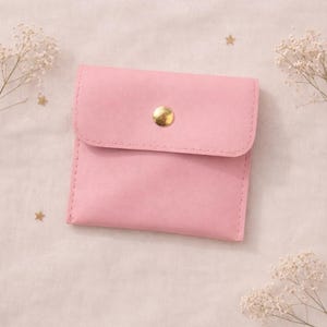 May include: A small, pink, square pouch with a flap closure and a gold-colored button. The pouch is made of a smooth material and has stitched edges. Decorative sprigs of white flowers and gold stars are in the background.