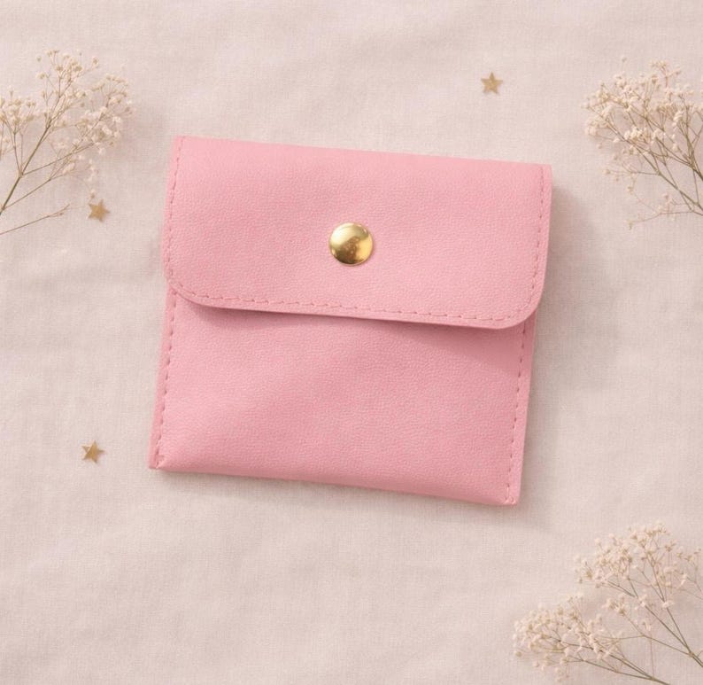 May include: A small, pink, square pouch with a flap closure and a gold-colored button. The pouch is made of a smooth material and has stitched edges. Decorative sprigs of white flowers and gold stars are in the background.