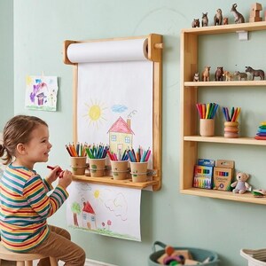 May include: A wall-mounted light wood paper roll dispenser with a drawing surface and four attached cups holding colored pencils, positioned next to a three-tiered light wood wall shelf containing various small animal figurines and art supplies.