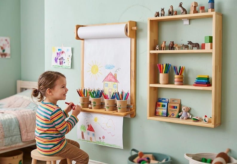 May include: A wall-mounted light wood paper roll dispenser with a drawing surface and four attached cups holding colored pencils, positioned next to a three-tiered light wood wall shelf containing various small animal figurines and art supplies.