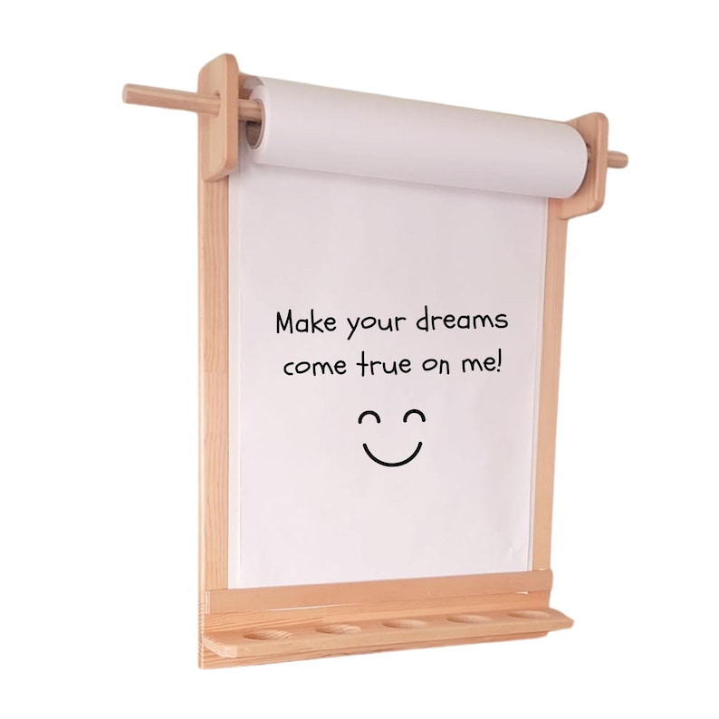 May include: A wall-mounted light wood paper roll dispenser featuring a horizontal rod, a flat backing board, and a bottom shelf with five circular cutouts. The white paper displays the black text "Make your dreams come true on me!" and a smiley face.