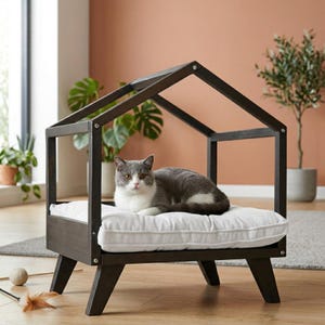 May include: A dark brown cat bed with a house-shaped frame. The bed has a white cushion and is elevated on angled legs. A grey and white cat is resting on the cushion.