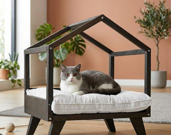 May include: A dark brown cat bed with a house-shaped frame. The bed has a white cushion and is elevated on angled legs. A gray and white cat is resting on the cushion.