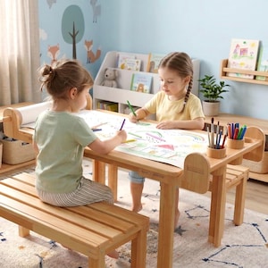 Op de afbeelding: Een lichtgekleurde houten activiteitentafel met een ingebouwde papierrolhouder en twee geïntegreerde cilindrische potloodhouders. De tafel heeft een glad rechthoekig oppervlak en wordt vergezeld door een bijpassende houten bank om op te zitten.